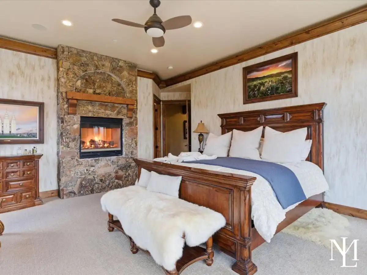 Spacious primary bedroom with carved wood bed, stone fireplace, soft neutral finishes, and cozy upscale mountain-lodge styling.