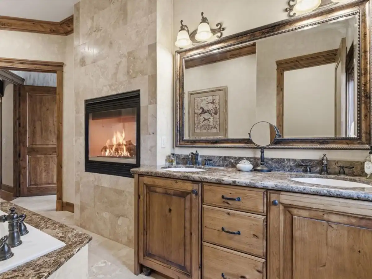 Primary bath vanity with dual sinks, wood cabinetry, stone countertops, framed mirror, and fireplace set into tiled wall.