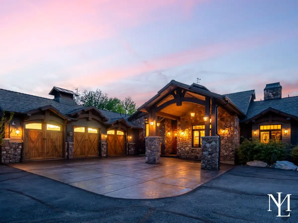 Luxury stone-and-timber mountain home in Huntsville, UT, at dusk with glowing entry, covered porte cochere, and three rustic wood garage doors.