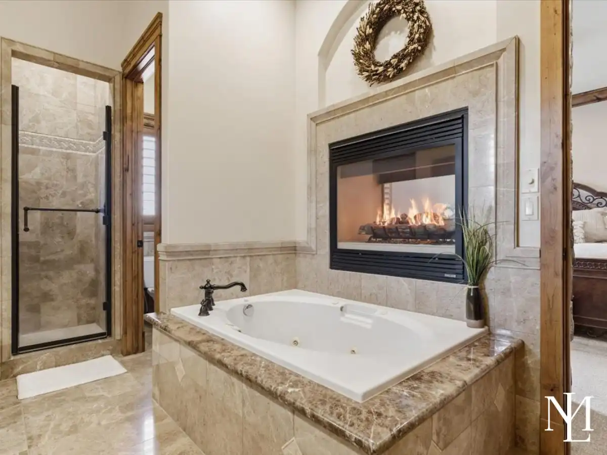 Spa-like primary bathroom with jetted soaking tub, walk-in shower, stone tile finishes, and a fireplace beside the bath.