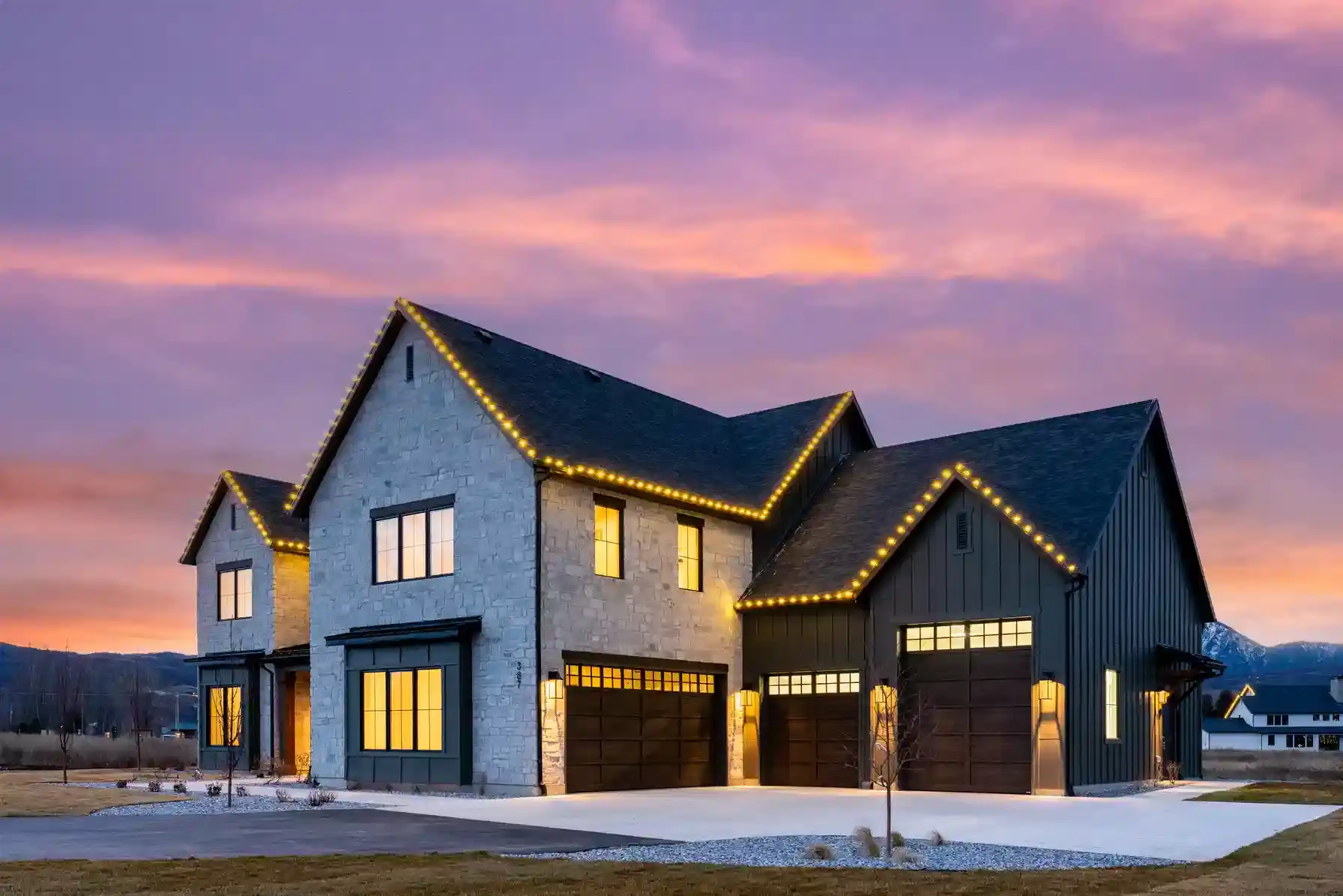 Luxury modern farmhouse with stone and dark siding, warm interior lighting, oversized garage doors, and roofline accent lights under a colorful twilight sky.