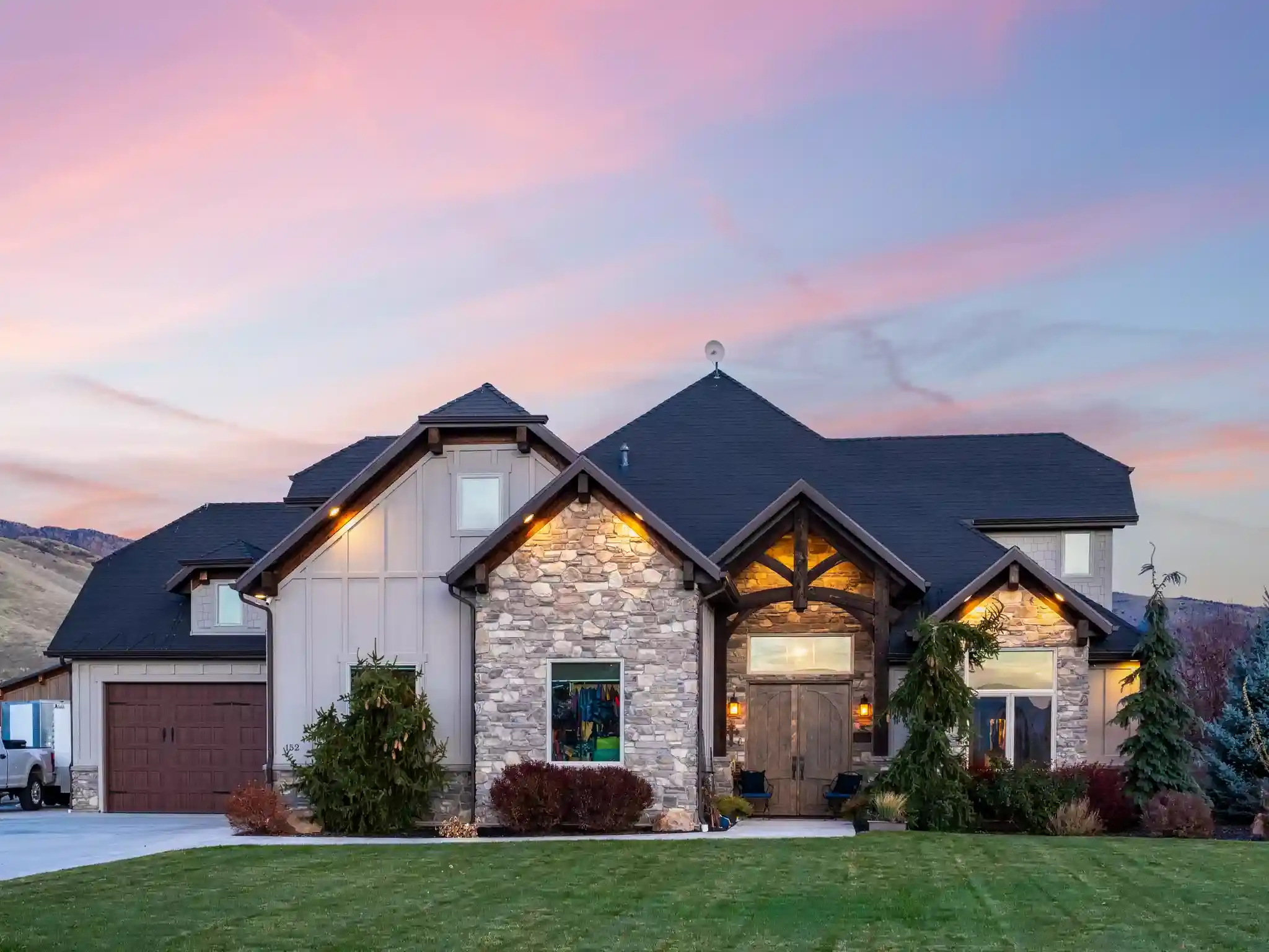 Custom luxury home with stone façade, vaulted timber entry, and manicured front lawn, photographed at twilight with soft mountain views in the background.