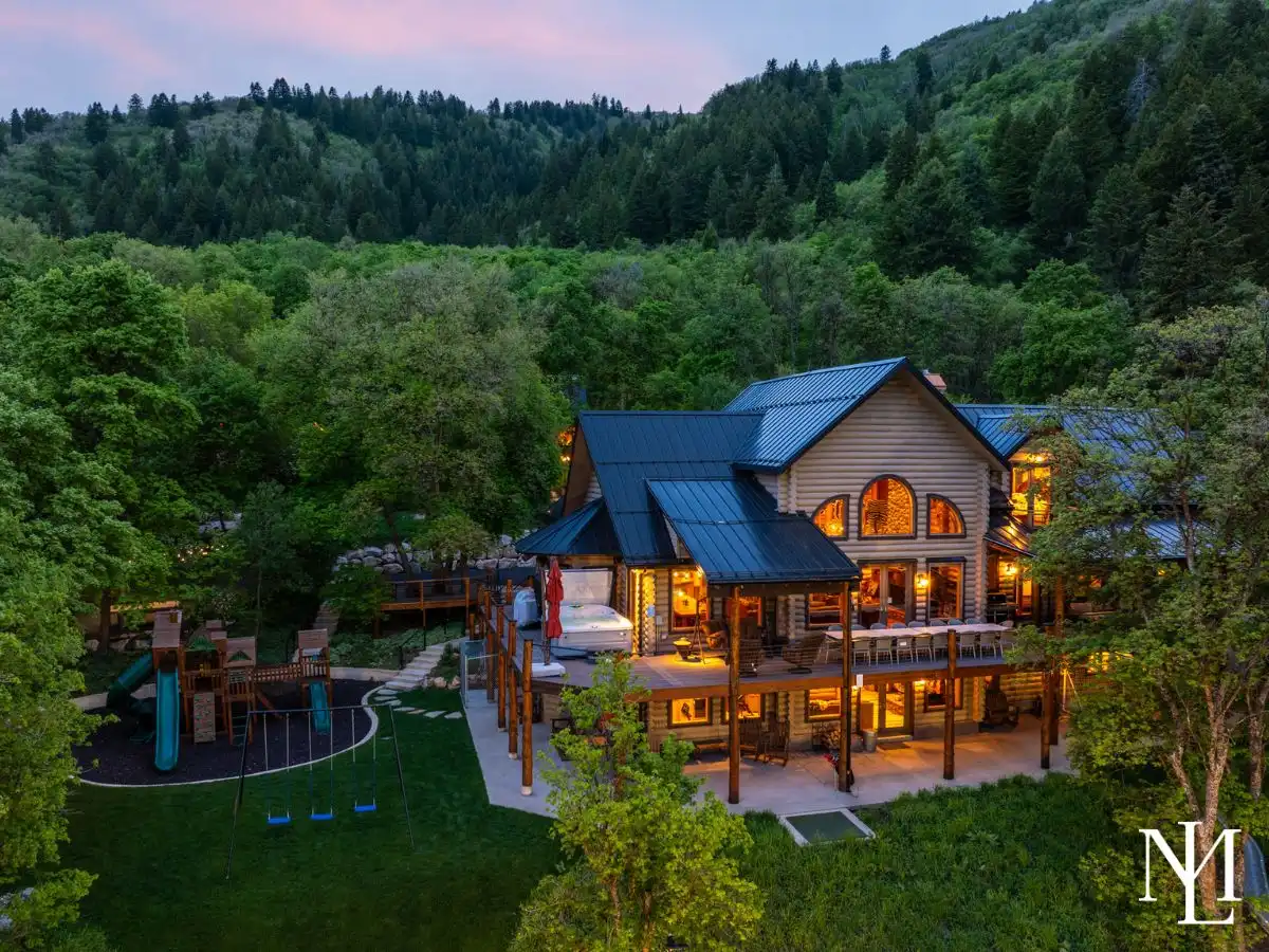 Twilight aerial view of a luxury log cabin estate with decks, hot tub, playground, and forested mountain backdrop.