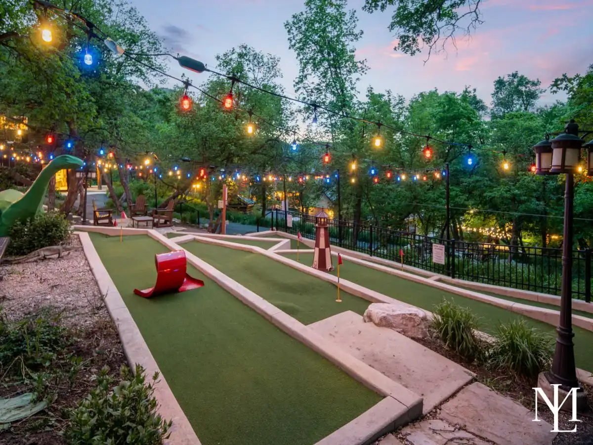 Outdoor dinosaur-themed mini golf course with string lights, putting greens, and wooded mountain setting.