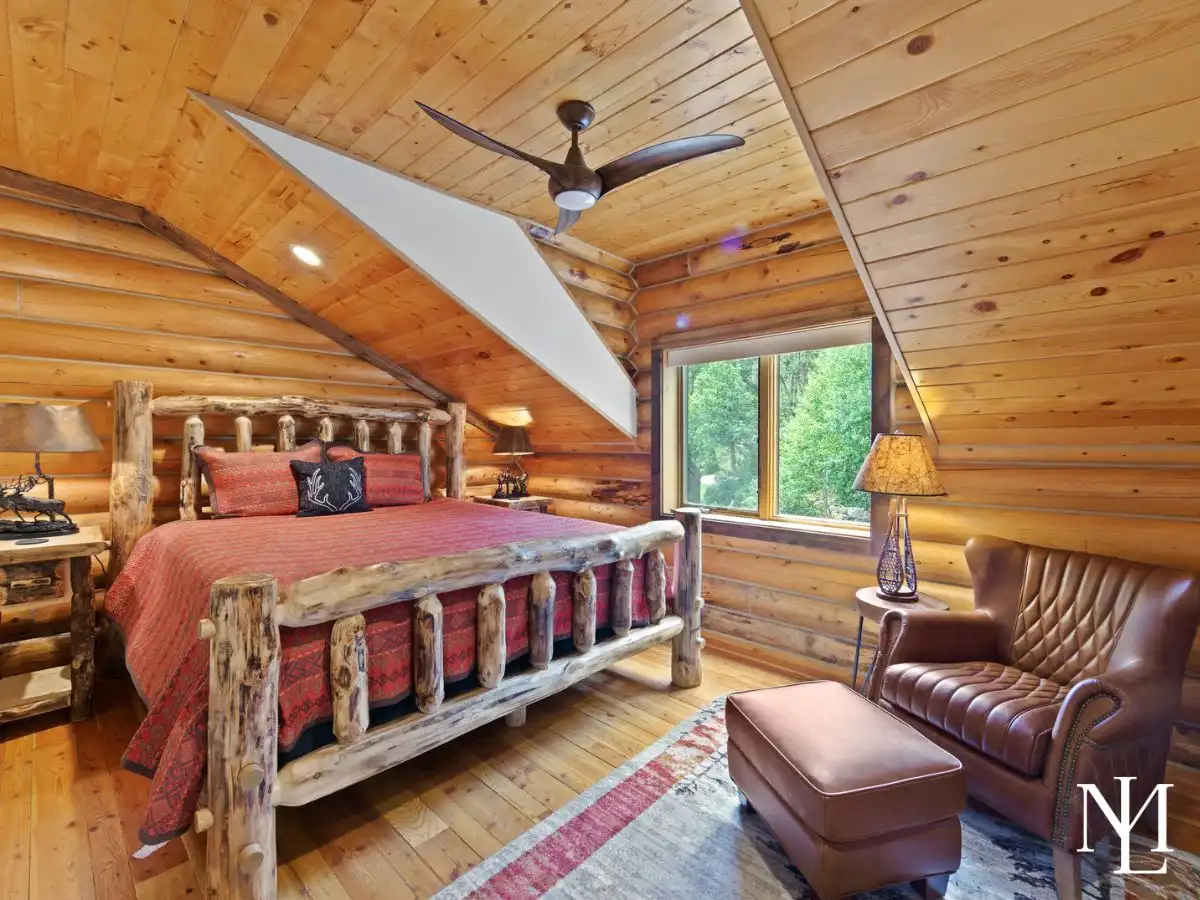 Cozy log cabin bedroom with rustic wood bed frame, vaulted wood ceiling, and forest views from the window.