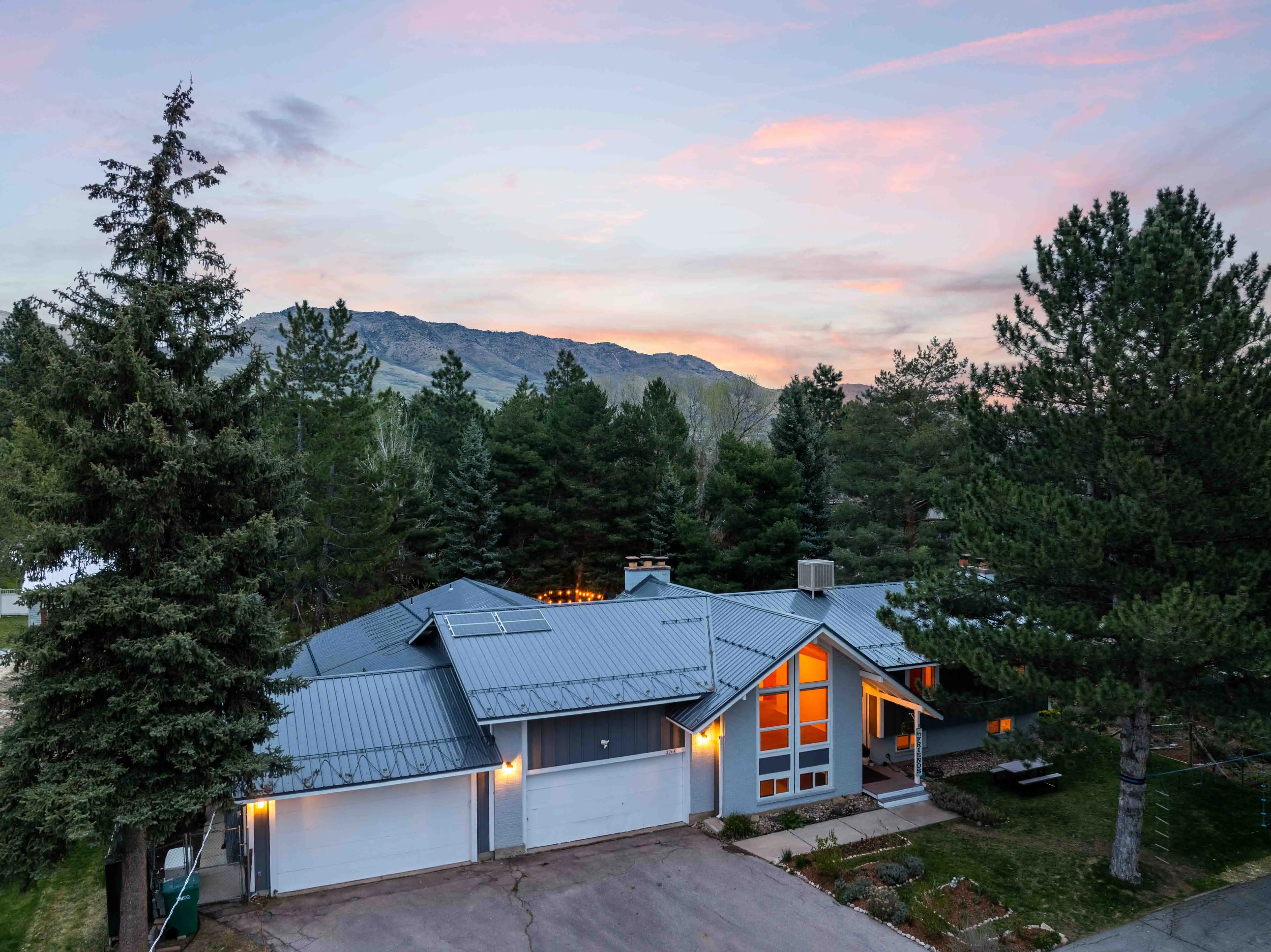 Twilight exterior of private Eden, Utah mountain home with metal roof, glowing windows, mature trees, and scenic mountain views.