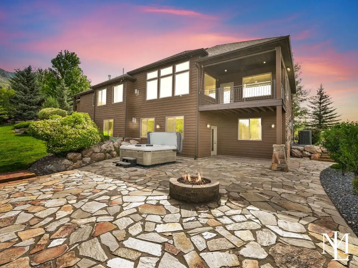 Rear exterior of Elkhorn hillside home in Eden Utah with covered deck, stone patio, hot tub, and fire pit