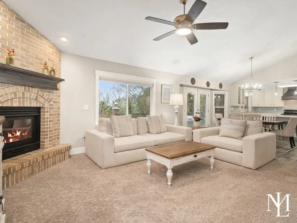 Main living room with brick fireplace, large picture window, and open layout to kitchen and dining area.