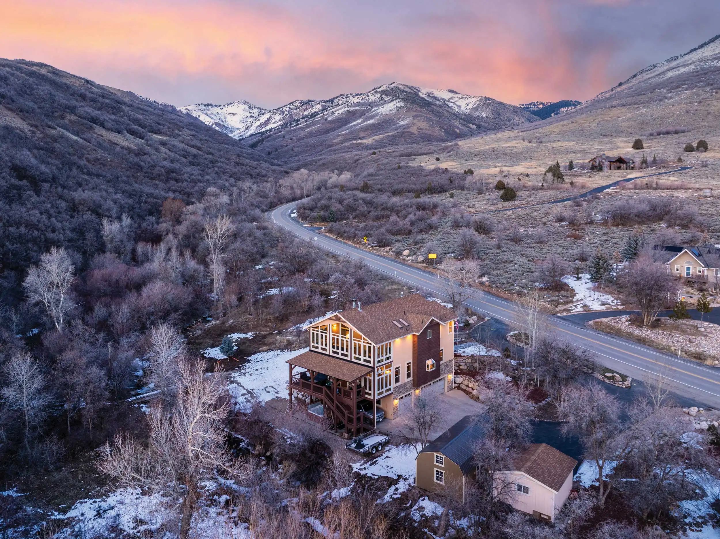 Aerial view of a mountain home with wraparound decks, outbuilding, creekside setting, and snow-dusted landscape beneath a colorful sunset sky.
