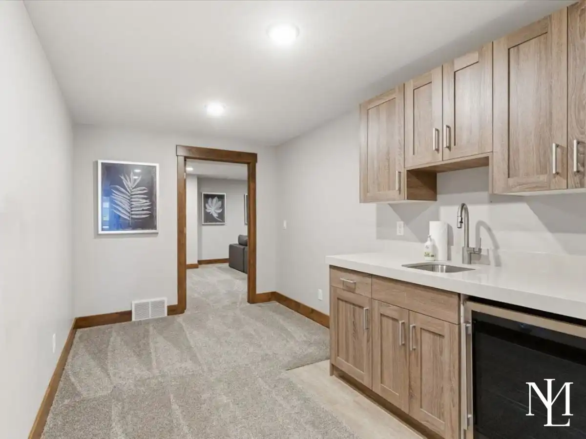 Lower-level kitchenette with wood cabinetry, sink, beverage fridge, and hallway access.