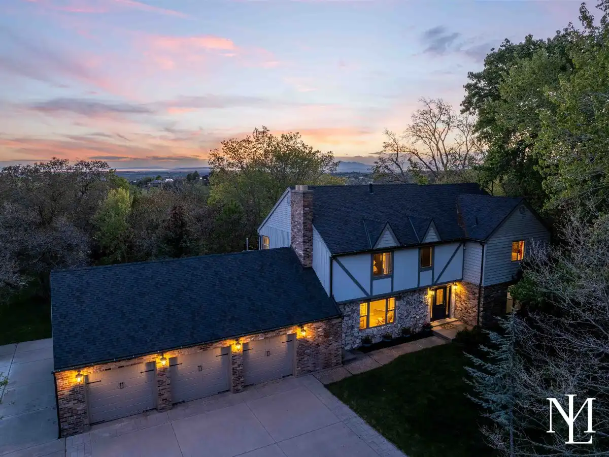 Twilight aerial view of a Tudor-style home in Uintah, Utah with a three-car garage, expansive driveway, mature trees, and warm interior lighting beneath a colorful sunset sky.