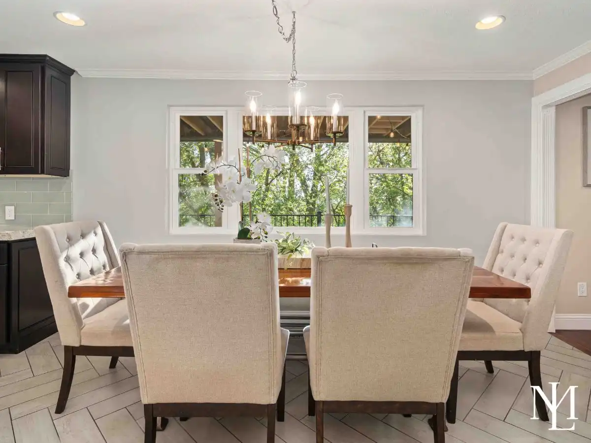 Dining area with upholstered chairs, modern chandelier, and large windows bringing in natural light and tree-filled views.