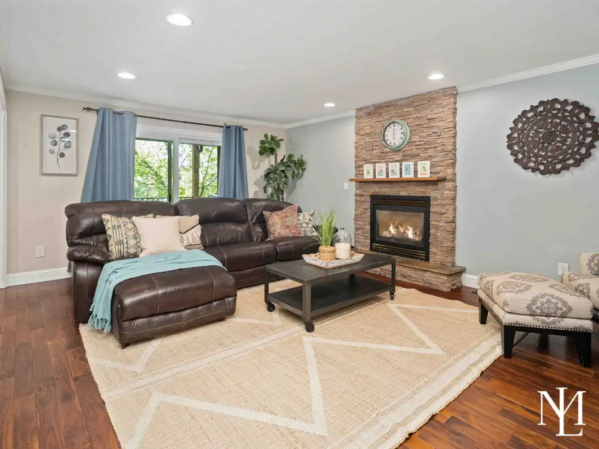 Living room with rich hardwood floors, a leather sectional, stacked-stone fireplace, soft gray walls, and large windows overlooking mature trees.