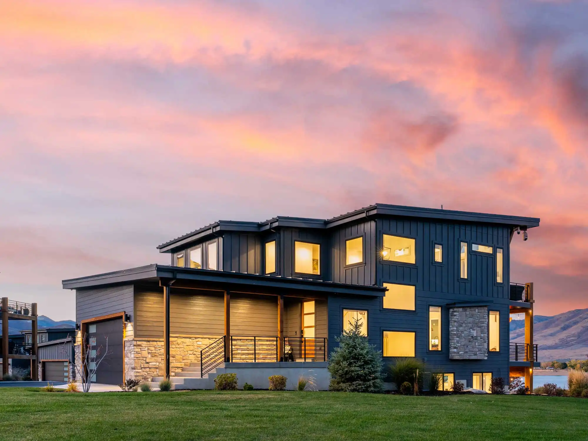 Modern luxury mountain home at sunset with dark exterior siding, stone accents, expansive windows, manicured lawn, mountain views, and lake access..