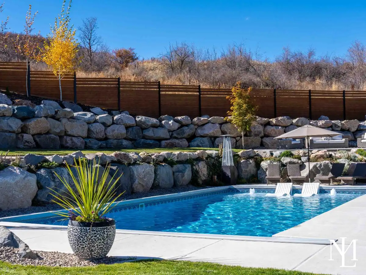 In-ground swimming pool with tanning ledge, lounge seating, terraced stone landscaping, and hillside privacy fence.
