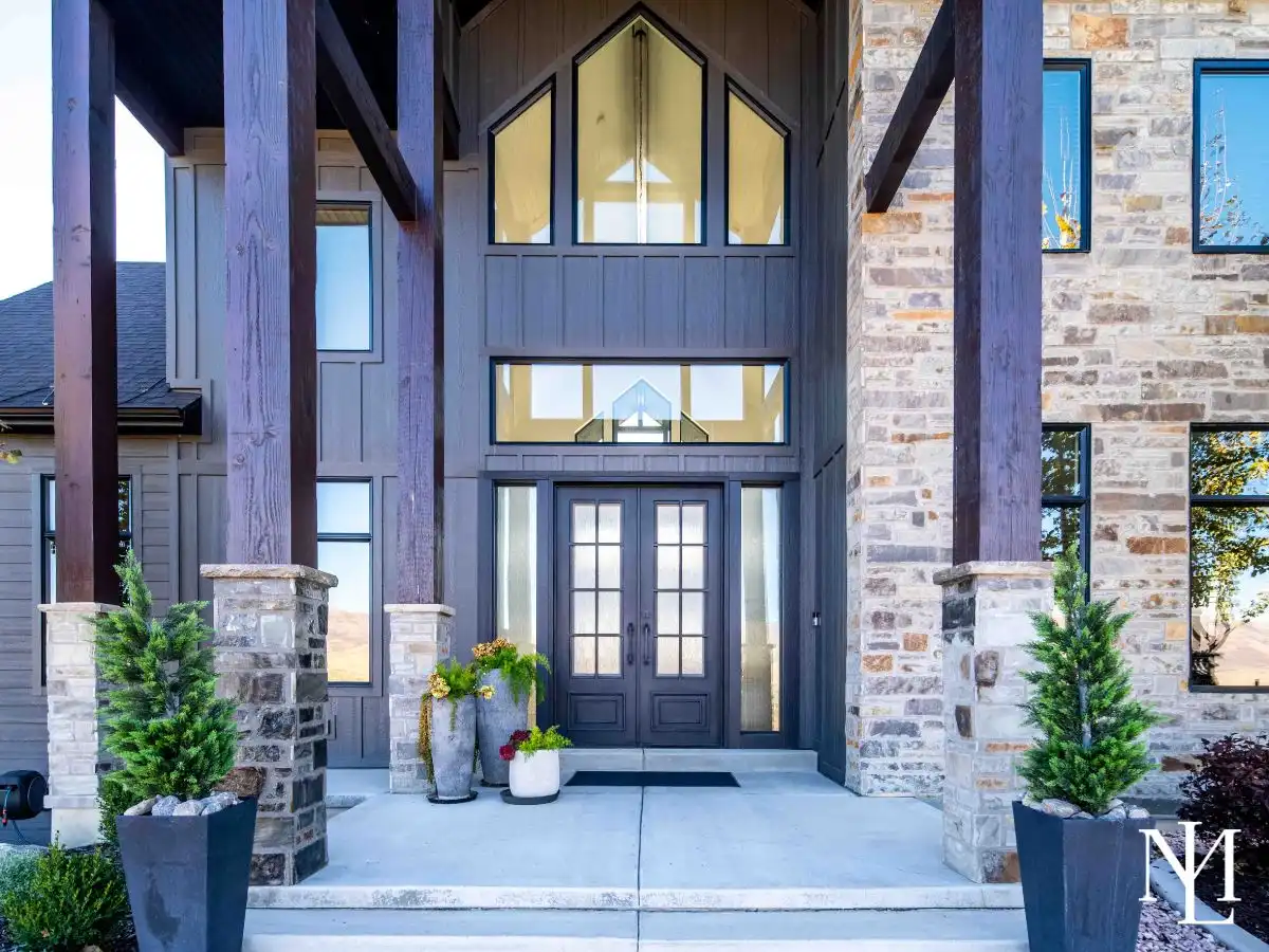 Front entry of modern mountain home with timber beams, stone columns, and double glass doors.