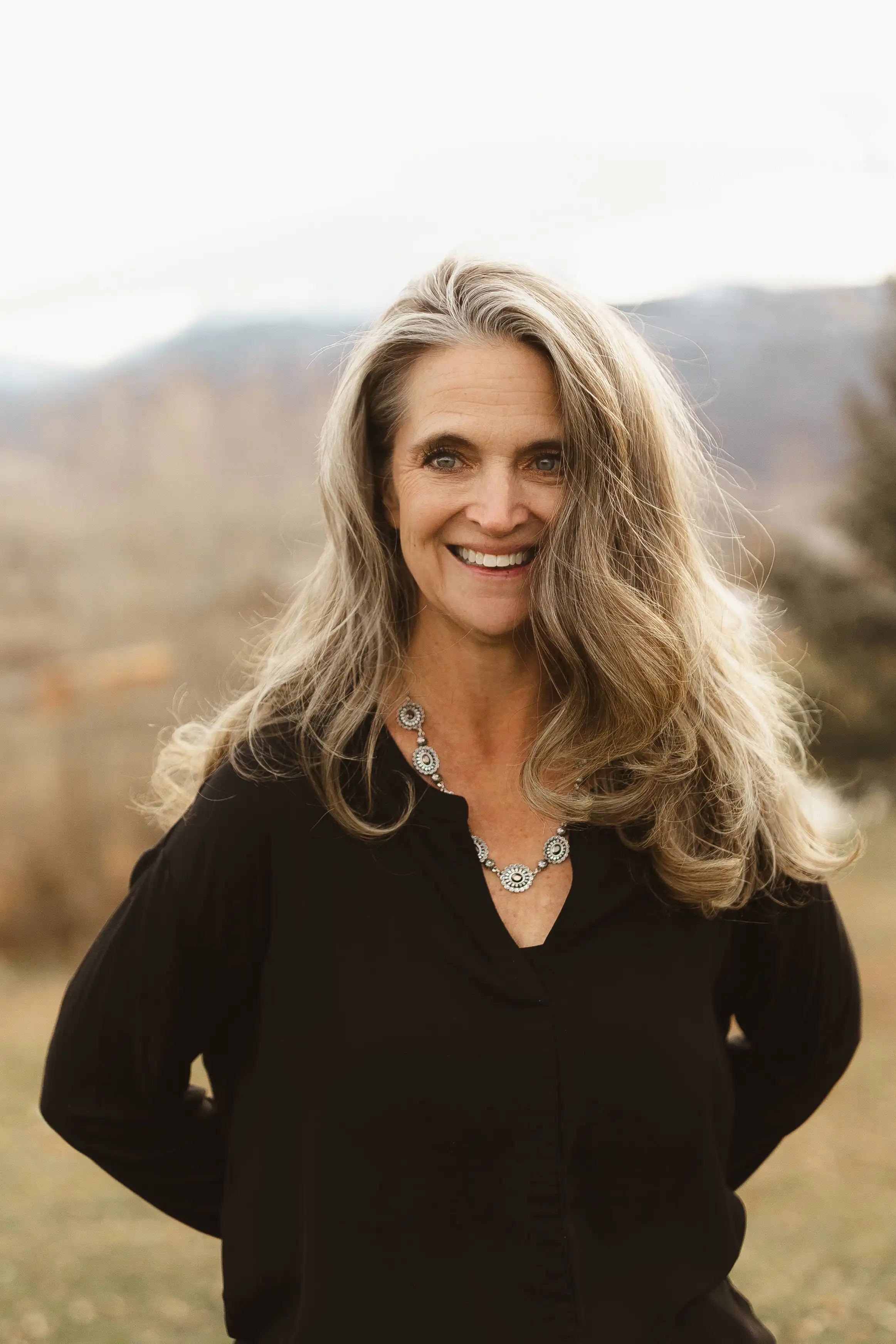 Outdoor professional portrait of Jennifer Stickler with long blonde hair wearing a black blouse and silver necklace, with soft mountain scenery in the background.