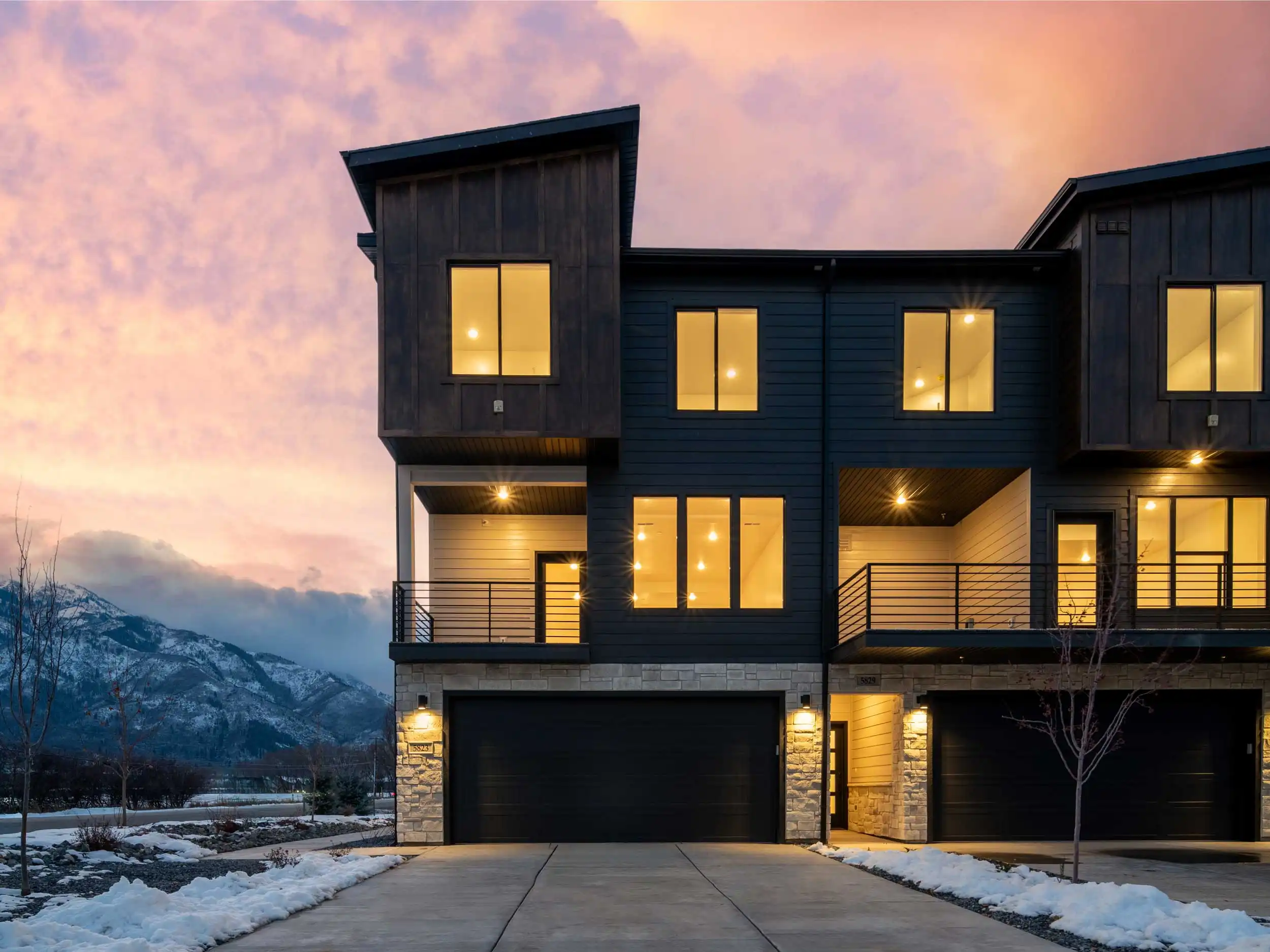 Modern Peaks townhome in Mountain Green with dark exterior, stone accents, illuminated windows, attached garage, snowy landscape, and mountain views at sunset.