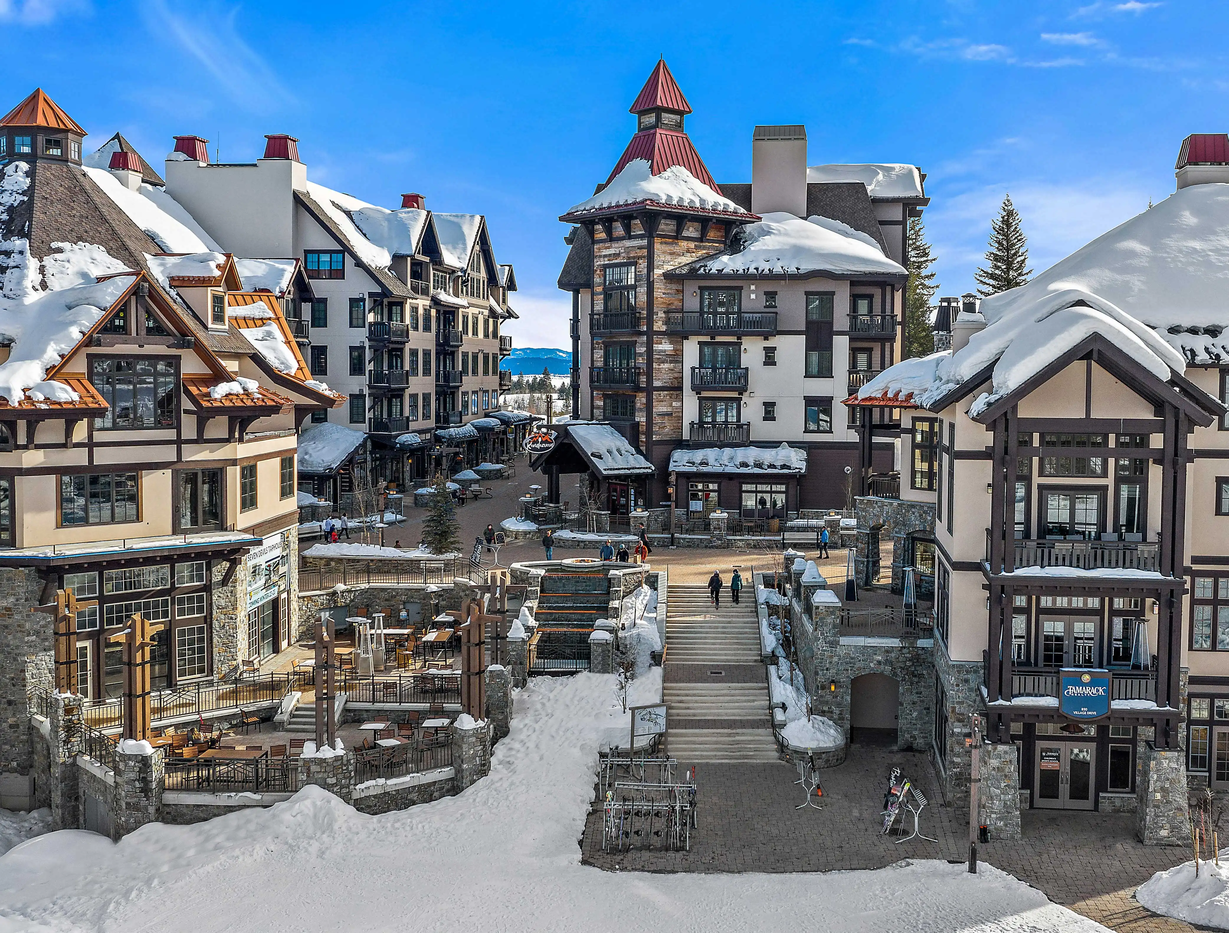 Snow-covered Village at Tamarack Resort with lodge-style buildings, outdoor plaza seating, and ski racks under a bright blue sky.