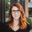Whit Sterner smiles in a professional outdoor headshot, wearing glasses and a black top with red hair styled over her shoulders.