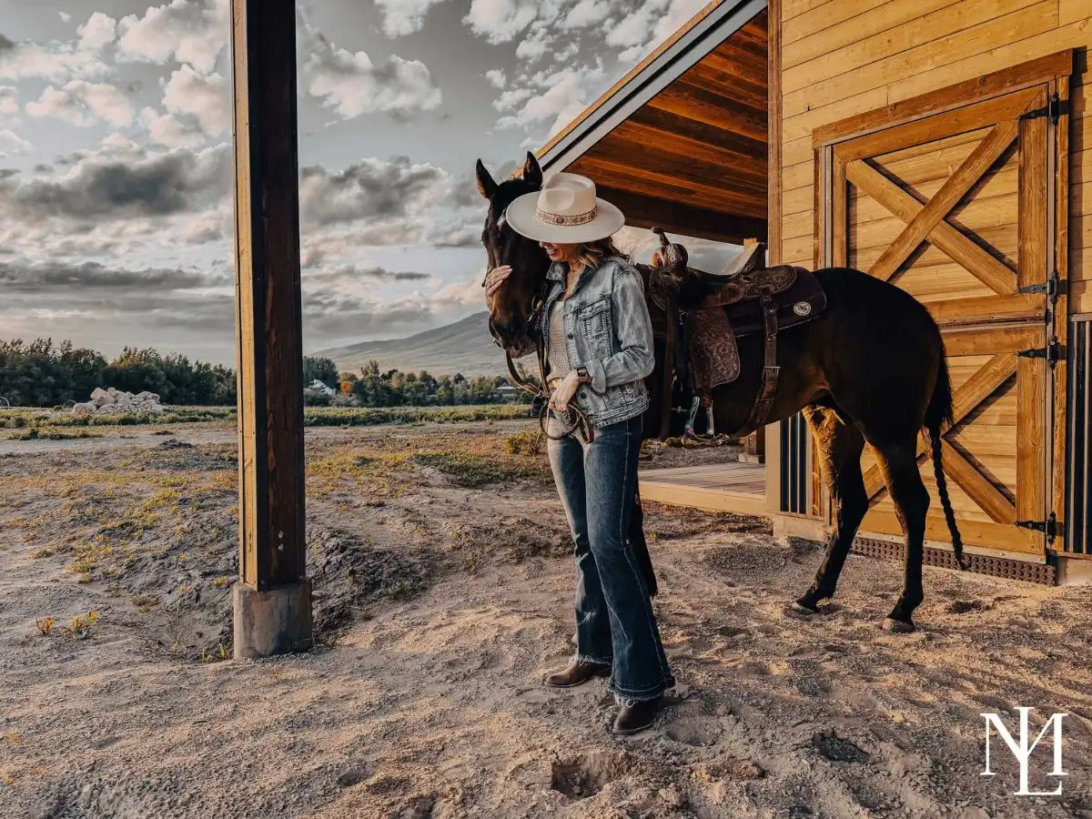Woman in a cowboy hat beside a tacked up horse next to a barn at sunset with mountain views.