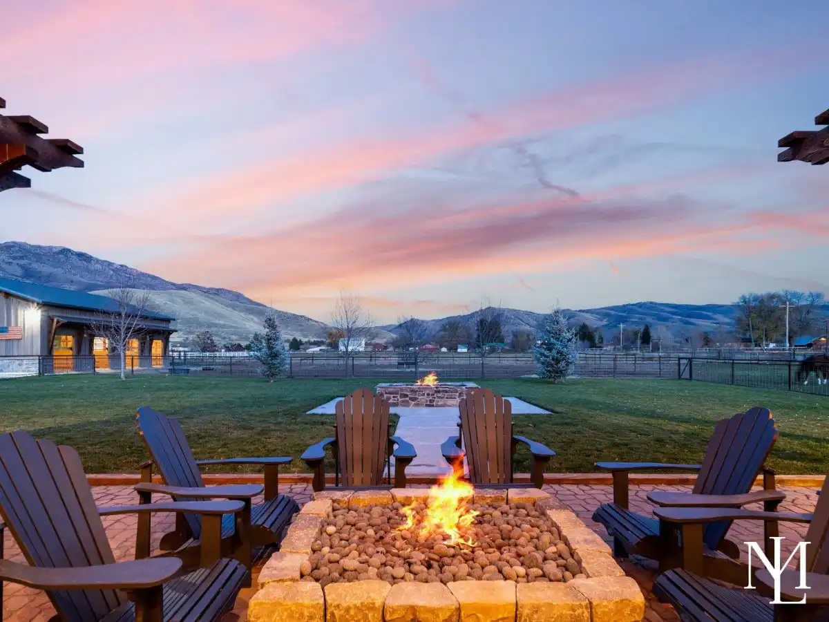 Backyard fire pit with Adirondack chairs overlooking fenced pasture and a barn under a colorful sunset sky.