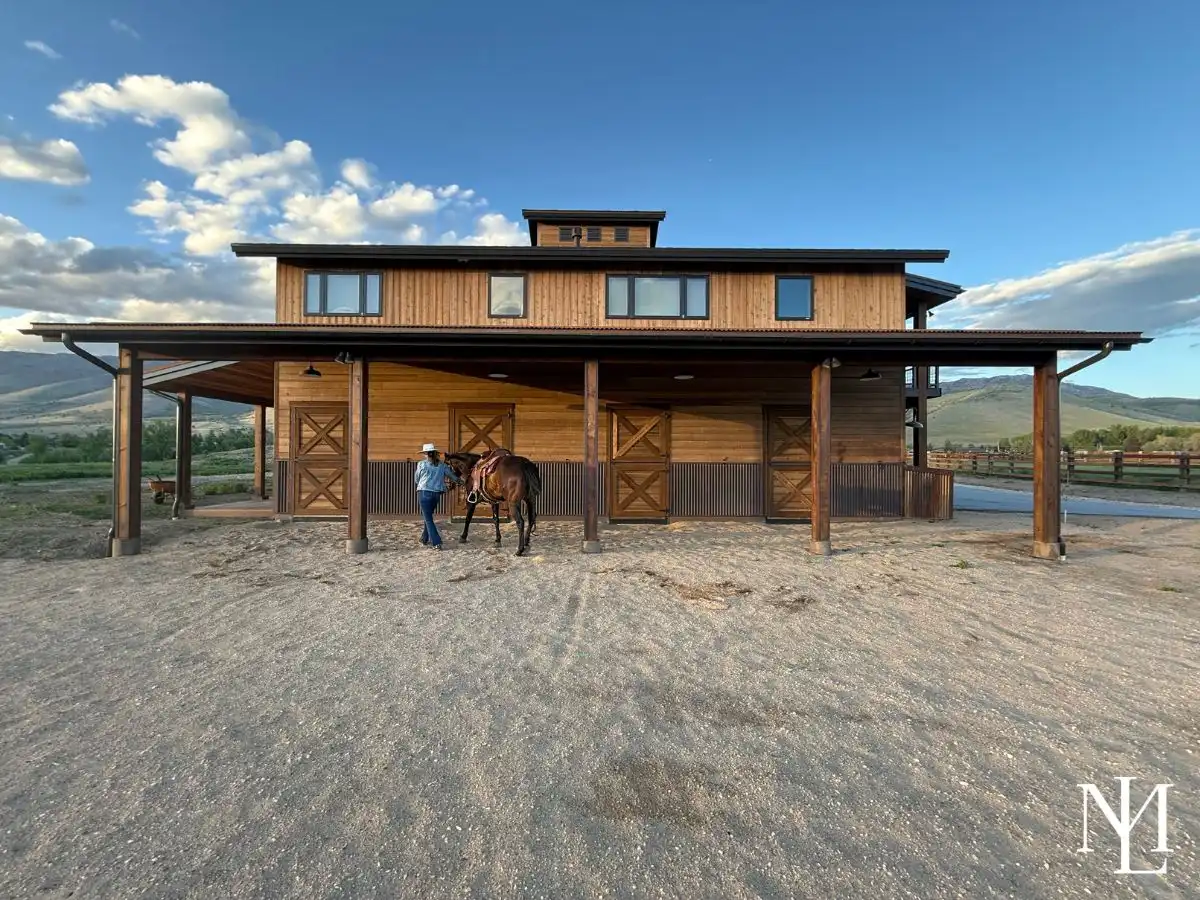 Wide exterior view of a modern wood barn with covered breezeway and a rider leading a horse, with mountains in the background.
