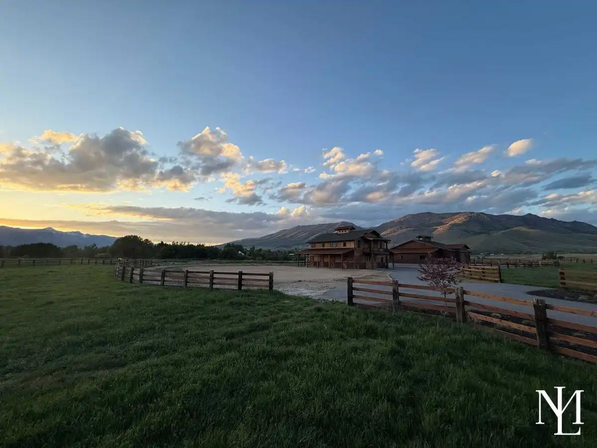 Wide view of a fenced horse property with barn, turnout area, and mountain backdrop during golden hour.