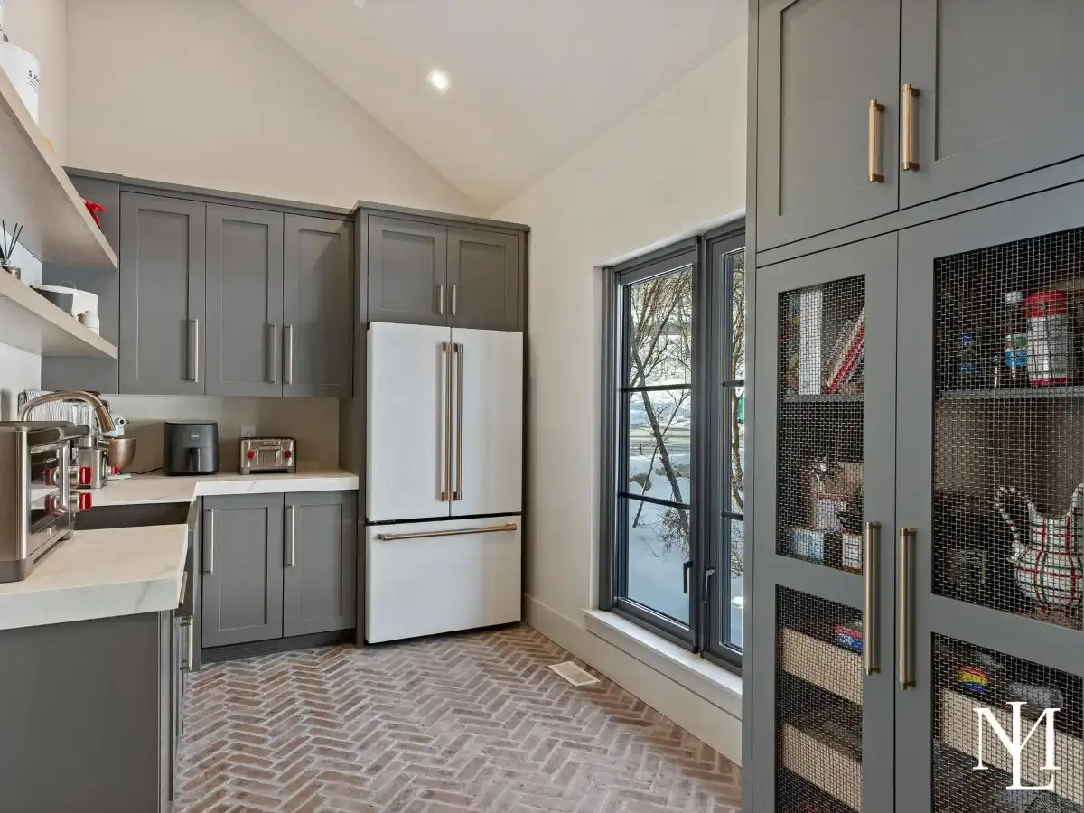 Luxury butler’s pantry with gray shaker cabinetry, white countertops, brick herringbone flooring, built-in storage, and large black-trimmed windows.