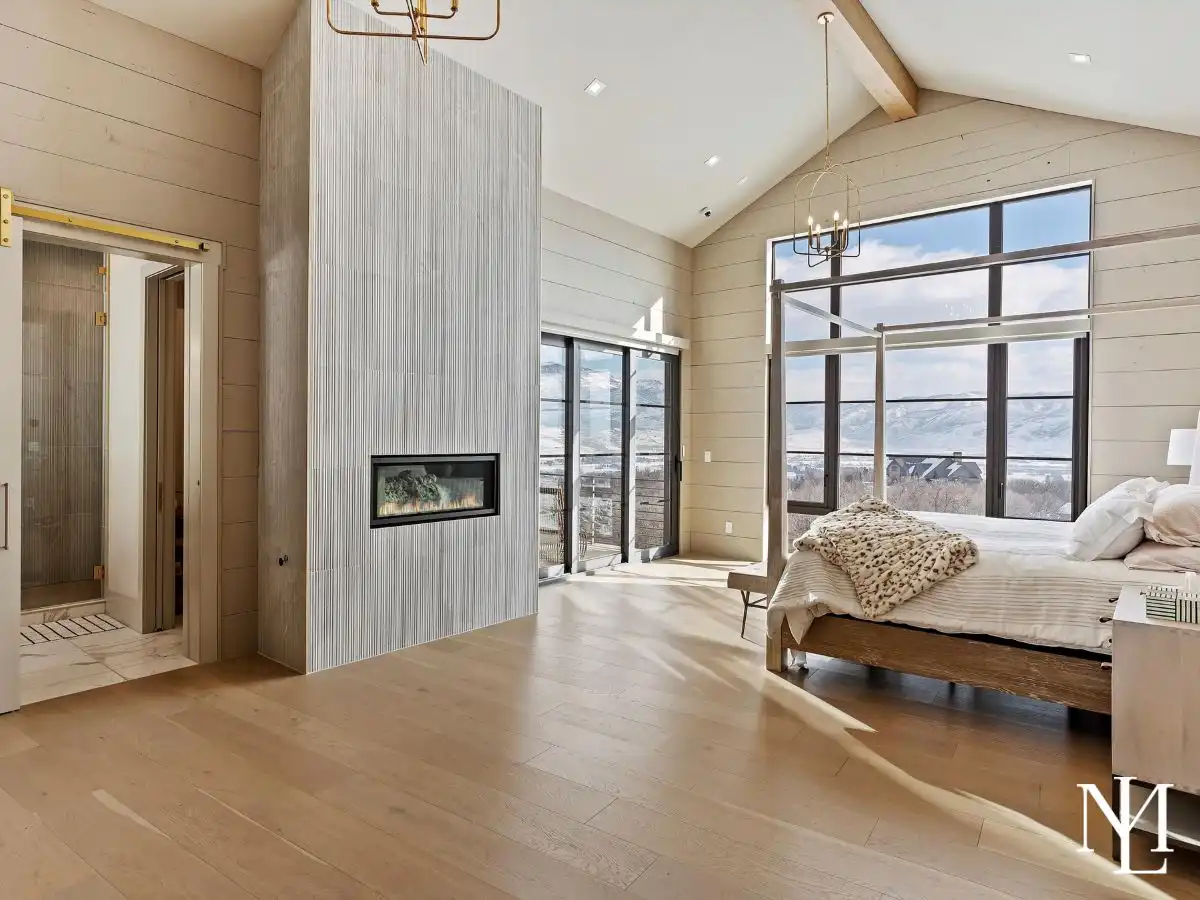 Luxury primary bedroom with fireplace feature wall, vaulted ceiling, expansive windows, and scenic mountain views.