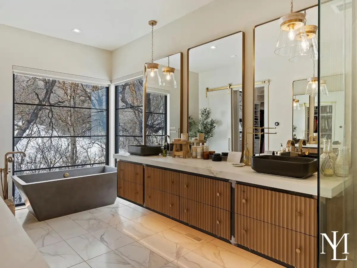 Spa-style primary bathroom with freestanding soaking tub, dual vanities, marble-look flooring, brass fixtures, and large windows overlooking snowy trees.