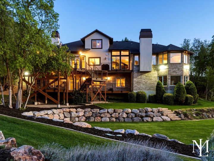 Rear exterior of upper bench Layton home with tiered landscaping, expansive deck, large windows, and private backyard setting at dusk.