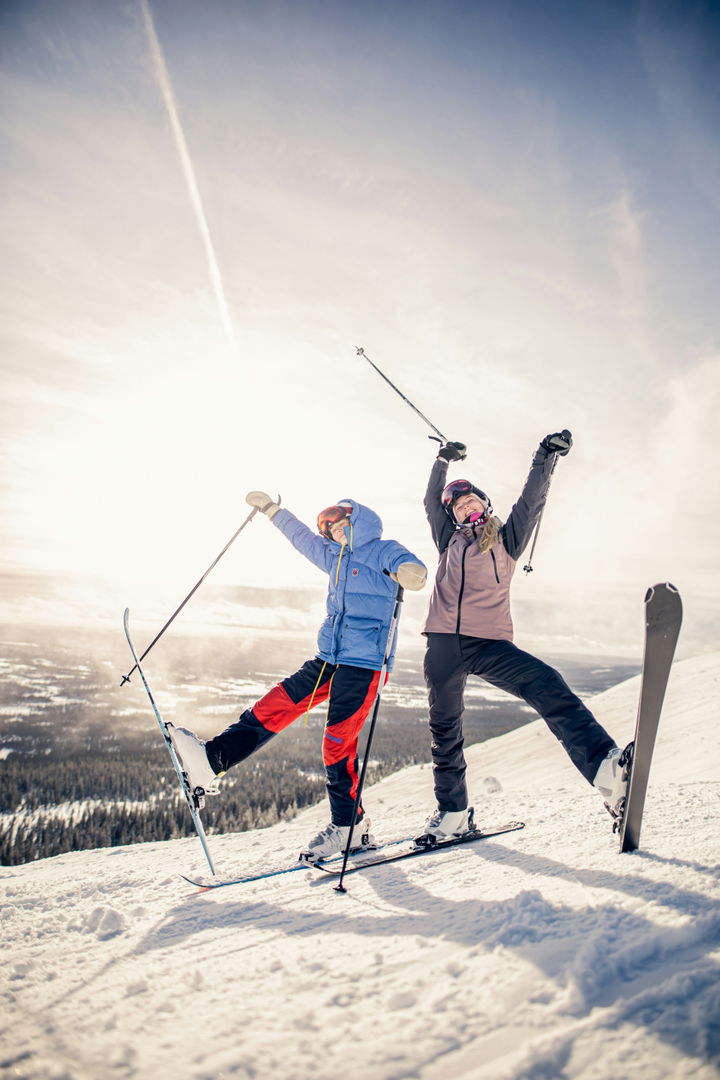 Two skiers celebrating on a sunny mountain slope with snowy terrain in the background.