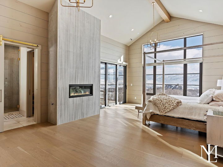 Luxury primary bedroom with fireplace feature wall, vaulted ceiling, expansive windows, and scenic mountain views.